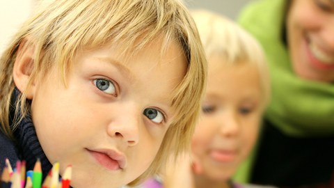 Ein blondes Kind schaut von links in die Kamera. Im Vordergrund Buntstifte, im Hintergrund ein weiteres Kind und eine Erwachsene, die sich über dessen Schulter beugt.