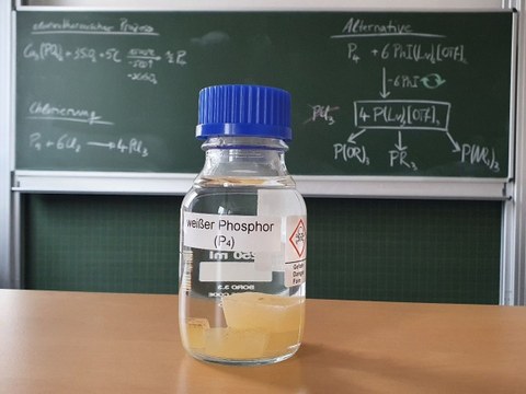 Eine Flasche mit weißem Phosphor steht auf einem Tisch vor einer beschriebenen Tafel.