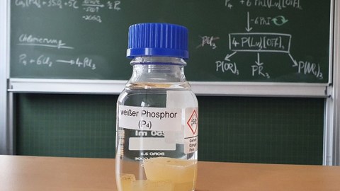 Eine Flasche mit weißem Phosphor steht auf einem Tisch vor einer beschriebenen Tafel.