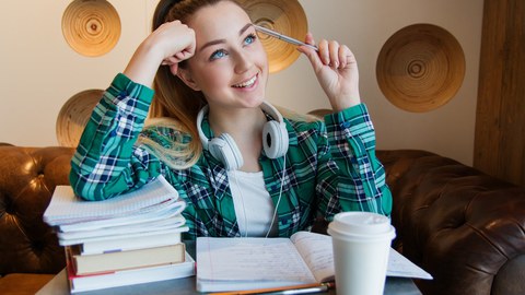 Eine junge Frau sitzt vor zwei Stapeln mit Büchern, hat den Kopf auf die eine Hand gestützt und hält sich mit der anderen einen Kuli an den Kopf