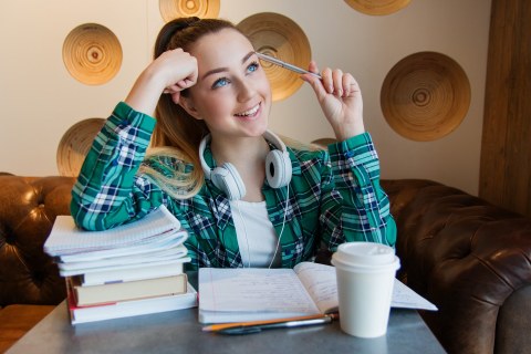 Eine junge Frau sitzt vor zwei Stapeln mit Büchern, hat den Kopf auf die eine Hand gestützt und hält sich mit der anderen einen Kuli an den Kopf