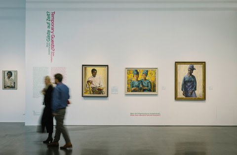 Foto aus der Ausstellung "Revolutionary Romances?" - zwei Personen bewegen sich vor einer Wand mit u.a. der Aufschrift "Gäste auf Zeit?/Temporary Guests?" und drei Gemälden, die unterschiedliche schwarze Personen zeigen.