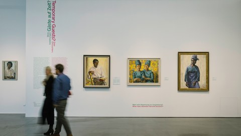 Foto aus der Ausstellung "Revolutionary Romances?" - zwei Personen bewegen sich vor einer Wand mit u.a. der Aufschrift "Gäste auf Zeit?/Temporary Guests?" und drei Gemälden, die unterschiedliche schwarze Personen zeigen.