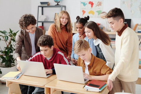 Um einen Tisch mit aufgeklappten Laptops stehen mehrere Schülerinnen und Schüler