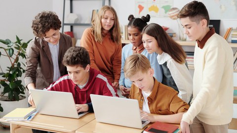 Um einen Tisch mit aufgeklappten Laptops stehen mehrere Schülerinnen und Schüler