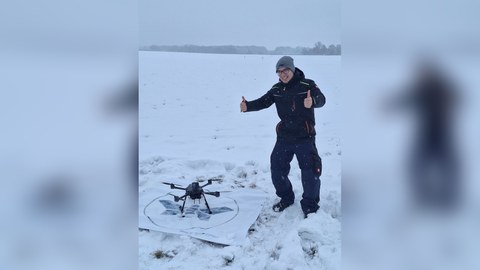 Junger Mann steht auf einer verschneiten Wiese, den rechten Daumen nach oben, im Bild rechts eine Drohne.