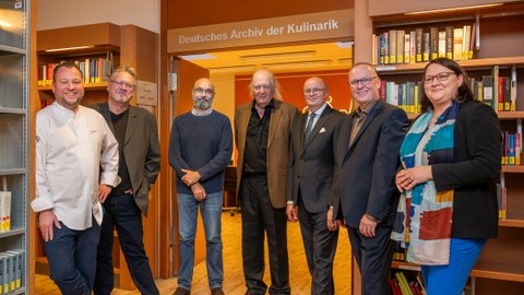 6 Männer und eine Frau stehen in einer Bibliothek um einen Durchgang herum. Über dem Durchgang der Schriftzug "Deutsches Archiv der Kulinarik"