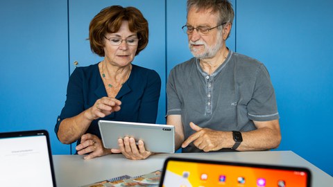 Ein älterer Herr hält ein Tablett in der Hand, eine ältere Dame sitzt neben ihm. Beide schauen auf das Tablett