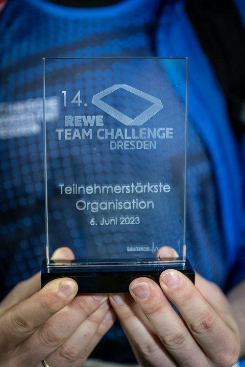 Zwei Hände halten eine Tafel in der Hand mit der Aufschrift "14. Rewe Team Challange Dresden. Teilnehmerstärkste Organisation. 6. Juni 2023