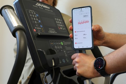 Display eines Ergometers, davor wird ein Smartphone mit einer geöffneten App ins Bild gehalten.