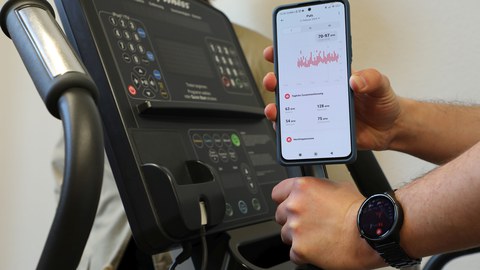 Display eines Ergometers, davor wird ein Smartphone mit einer geöffneten App ins Bild gehalten.