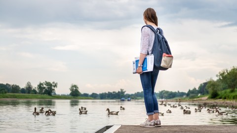 Eine junge Frau steht am Rande eines Stegs an der Elbe. Im Wasser schwimmen Enten. Die Frau hält Bücher unter dem Arm und trägt einen Rucksack.