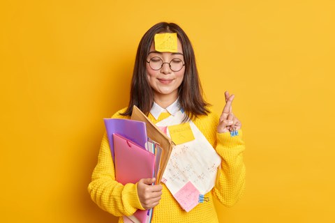 Junge Frau hält im rechten Arm viele Hefter und Umschläge, die linke Hand ist nach über gebeugt, die Finger gekreuzt. Auf der Stirn klebt ein PostIt.