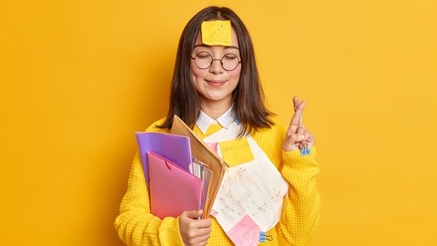 Junge Frau hält im rechten Arm viele Hefter und Umschläge, die linke Hand ist nach über gebeugt, die Finger gekreuzt. Auf der Stirn klebt ein PostIt.