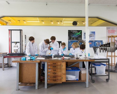 Eine Gruppe von Schüler:innen in weißen Laborkitteln schaut auf einen Tisch