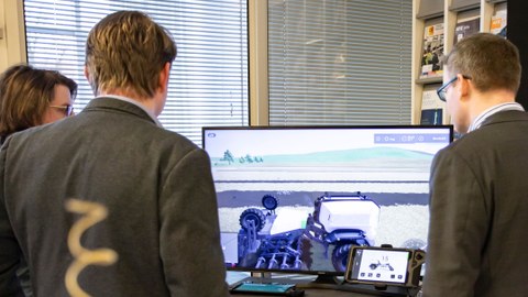 Drei Männer schauen auf einen Monitor, der die Visualisierung einer Landmaschine zeigt