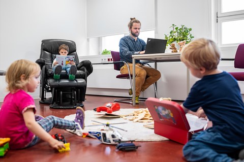 Drei kleine Kinder spielen, überall ist Spielzeug vereilt. Im Hintergrund arbeitet ein Mann am Laptop.