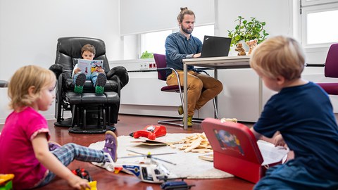 Drei kleine Kinder spielen, überall ist Spielzeug vereilt. Im Hintergrund arbeitet ein Mann am Laptop.