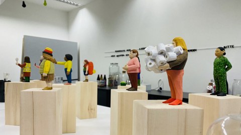 Eine Reihe bemalter Holzfiguren auf Holzsockeln, die unterschiedliche Charaktere vom "Hutbürger" bis Angela Merkel darstellen.