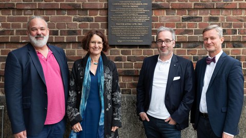 Gruppenfoto (eine Frau, drei Männer) vor einer Ziegelwand. Daran eine Gedenktafel "Historische Stätte der Chemie"