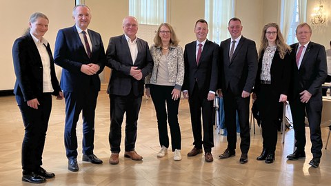 Gruppenfoto mit fünf Männern in dunklen Anzügen und drei Frauen mit hellen Blusen und dunklen Hosen