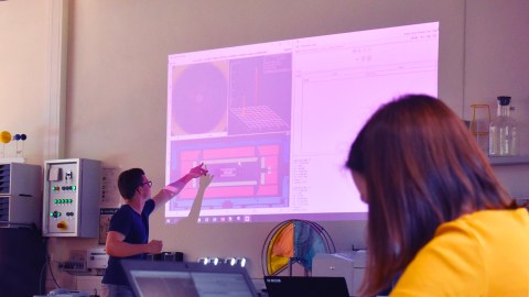 Im Hintergrund steht ein junger Mann vor einem Beamerbild und zeigt auf eine Grafik. Im Vordergrund rechts sitzt eine Person am Laptop.