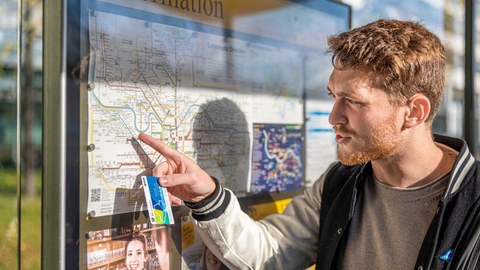 Junger Mann mit einer Campuscard in der Hand steht vor einem Verkehrsplan der Dresdner Verkehrsbetriebe.