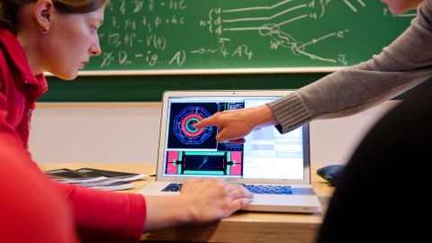 Zwei Personen arbeiten an einem Laptop, im Hintergrund eine mit physikalischen Formeln beschriebene Tafel.