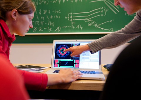 Zwei Personen arbeiten an einem Laptop, im Hintergrund eine mit physikalischen Formeln beschriebene Tafel.