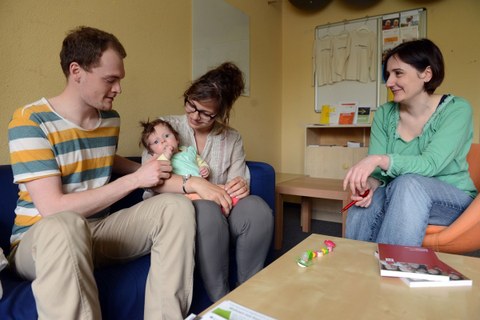 Eine junge Familie mit Baby sitzt auf einer Couch, gegenüber eine Beraterin.