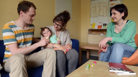 Eine junge Familie mit Baby sitzt auf einer Couch, gegenüber eine Beraterin.