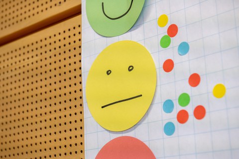 Drei Papier-Smileys übereinander an einer gelochten Wand. Der gelbe in der Mitte ist vollständig zu sehen, grün (darüber) und rot (darunter) nur als Ausschnitt.