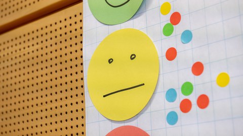 Drei Papier-Smileys übereinander an einer gelochten Wand. Der gelbe in der Mitte ist vollständig zu sehen, grün (darüber) und rot (darunter) nur als Ausschnitt.