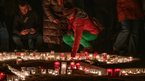 Im Vordergrund stehen viele Kerzen. Im Hintergrund zündet eine Frau eine Kerze an.