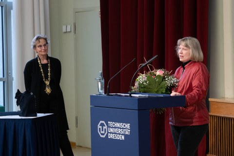 Rechts im Bild steht eine Frau am Rednerpult. Vor ihr liegt ein Blumenstrauß. Links im Hintergrund ist eine weitere Frau.