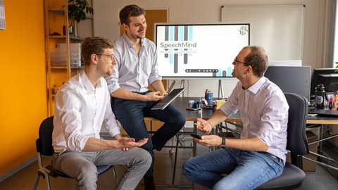 Drei junge Männer sitzen einander zugewandt in einem Büro und unterhalten sich.