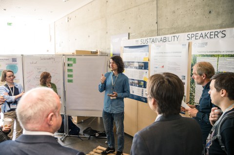 Fotoaufnahme einer Person, die vor Flipcharts und plakatierten Stellwänden steht und zu einer Gruppe an Personen spricht.