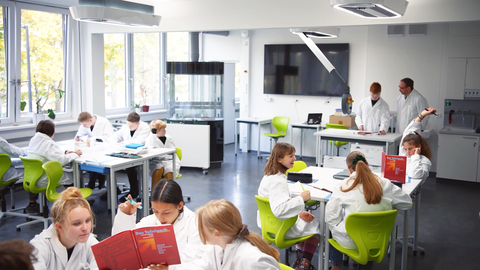 Fotoaufnahme eines Klassenraums, in dem die Schülerinnen und Schüler in Gruppen an Tischen beisammen sitzen und lernen.