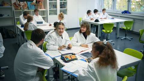 Fotoaufnahme eines Klassenraums, in dem die Schülerinnen und Schüler in Gruppen an Tischen beisammen sitzen und miteinander sprechen.