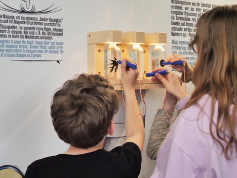 Fotoaufnahme von zwei Personen, die in einer Ausstellung mit einem stiftähnlichen Gegenstand an Glasbehältern entlangfahren.