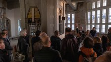 Mehrere Personen hören einem Mann zu vor einem Bauelement auf einer Baustelle in einem Gebäude.