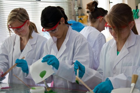 Mädchen mit Laborkitteln, Schutzbrille- und Handschuhen experimentieren mit Farbe, Pinseln und Papiertüchern.