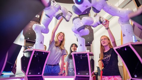 3 junge Frauen beobachten und begreifen vor ihnen aufgebauteRoboterarme.