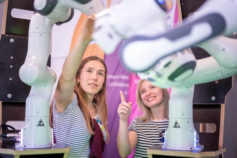 Foto von zwei jungen Frauen in schwarz-weiß gestreiften T-Shirts, die inmitten von weißen Roboterarmen stehen und diese betrachten.