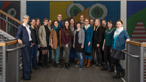 Foto einer Gruppe lächelnder Menschen, die im Halbkreis auf einem grauen Podest zwischen zwei Metalltreppen stehen.