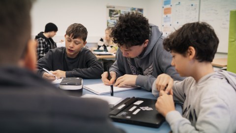 Drei Jungen sitzen an einem runden Tisch in einem Klassenzimmer. Der Junge links im Bild schreibt, der Junge in der Mitte auch. Vor dem Jungen rechts liegt ein Laptop.