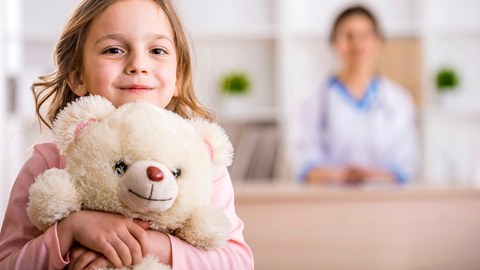 Ein junges Mädchen mit einem Teddybär im Arm links im Vordergrund, rechts im Hintergrund verschwommen eine Frau.