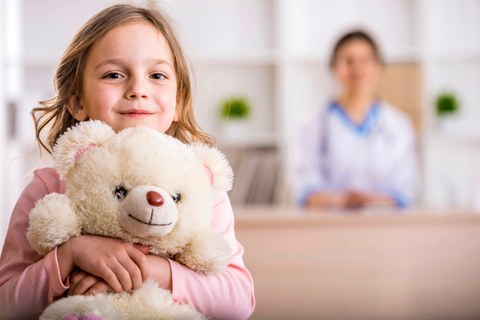 Ein junges Mädchen mit einem Teddybär im Arm links im Vordergrund, rechts im Hintergrund verschwommen eine Frau.
