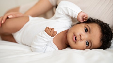 Baby mit dunkler Hautfarbe und schwarzen Haaren liegt seitlich auf einem weißen Tuch und schaut zum Betrachter. Auf seinem Bein eine Hand mit langen Fingernägeln.