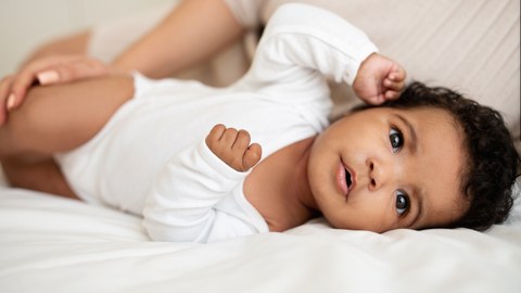 Baby mit dunkler Hautfarbe und schwarzen Haaren liegt seitlich auf einem weißen Tuch und schaut zum Betrachter. Auf seinem Bein eine Hand mit langen Fingernägeln.